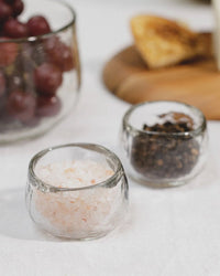 The Collective Pebbled Condiment Bowls - Set of 4 hold pink Himalayan salt and black peppercorns on a white tablecloth, with grapes and toast in the background—perfect for serving condiments at any gathering.