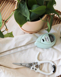 A potted plant with green leaves sits on a cloth beside a soil moisture meter and Jungle Club Pruning Shears in White. The items are arranged on a light surface, complemented by a woven mat in the background.