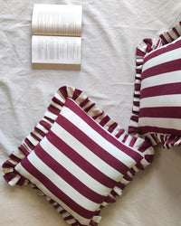 Two Casa Amarosa Cherry Ruffle 16x16in pillows with maroon and white stripes and ruffled edges rest on a light fabric surface beside an open book, creating a cozy, inviting scene.