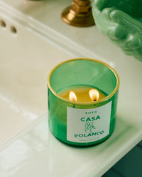 A green glass ROEN Casa Polanco Candle with two flames sits on a white countertop, its coconut soy wax blend scent filling the air beside a green bowl and a brass object in the background.