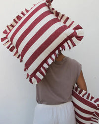 A person in a taupe shirt and white pants holds two Casa Amarosa Cherry Ruffle 16x16in pillows—one covering their face, the other by their side—against a plain white background.