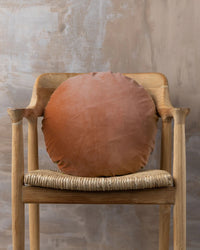 A wooden chair with a woven seat displays the Saarde Linen/Velvet Round Pillow with Feather Insert in Terracotta, set against a textured beige wall.