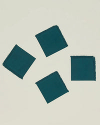 A set of four Hawkins New York Essential Cocktail Napkins in Peacock, made from 100% cotton with frayed edges, are displayed scattered diagonally on a light neutral background.