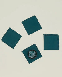 Four Hawkins New York Essential Cocktail Napkins in Peacock, made from 100% cotton with frayed edges, are spread on a light surface. One napkin holds a small bowl with green items, arranged loosely in a diagonal line.