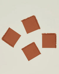 A set of four Hawkins New York Essential Cocktail Napkins in Terracotta, made from 100% cotton with fringed edges, is displayed on a plain, light-colored surface.