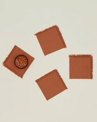 A set of four Hawkins New York Essential Cocktail Napkins in Terracotta, made from 100% cotton with frayed edges, are displayed on a light surface; one holds a glass of iced tea while the others are empty.