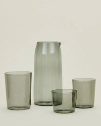 A set of Hawkins New York Essential Glassware—Pitcher in Smoke with a spout and three matching smoky gray tumblers of different heights, showcased on a light background.