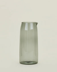 A tall, transparent 52 oz borosilicate glass pitcher in Smoke by Hawkins New York stands upright against a light beige background, featuring a small spout and exuding simplicity and modern elegance.