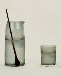 The Essential Glassware - Pitcher in Smoke from Hawkins New York, a frosted 52 oz borosilicate glass pitcher with wooden spoon, stands next to a matching water glass on a light surface with visible condensation.