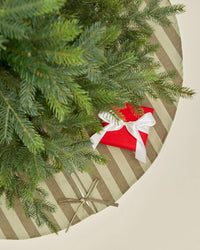 A top-down view of a Christmas tree on the Hawkins New York Essential Striped Tree Skirt in Olive/Sage, with a red gift wrapped with white ribbon and a smaller brown paper gift tied with twine beneath the branches.