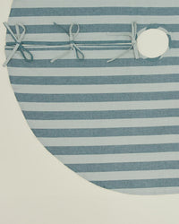 The Essential Striped Tree Skirt in Sky/Peacock by Hawkins New York is a circular cotton skirt with horizontal light blue and gray stripes, three light blue ribbon ties, and a round opening for easy placement around your tree trunk.