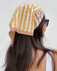 A woman with long brown hair models the Casa Amarosa Stripe Hand Block Printed Scarf in Yellow & Brown as a headscarf, styled with black sunglasses and a white sleeveless top against a plain light wall.