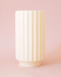 The Monroe Vase in Vintage White by Jungle Club, a tall cylindrical ceramic vase with vertical ridges and a modern minimalist design in soft cream, sits on a light pink background.
