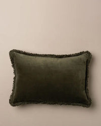 The Saarde Velvet Lumbar Pillow w/ Feather Insert in Dark Olive, featuring matching fringe on all sides and a plush feather down insert, is styled against a plain beige background.