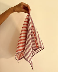 A hand displays Casa Amarosa's Stripe Hand Block Printed Scarves in purple and red against a plain beige wall.