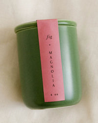 A green jar Fig & Magnolia Candle by LEIF, 4 oz. with a pink label and wooden wick, sits on a light-colored surface.