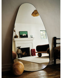 Ad At Home: Architectural Digest by Random House features an asymmetrical floor mirror that reflects a cozy room with a white fireplace, art, a bed, unique decor like wooden sculptures, and a round wooden object on the hardwood floor.