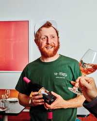 A smiling man with a red beard, wearing a green shirt and white cap, holds a bottle of Salad Pizza Wine by Random House. Another hand in the foreground raises a glass of rosé toward him, with red art hanging on the wall behind them.