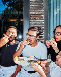 Four people enjoy Salad Pizza Wine by Random House outdoors near a brick wall and window—three wearing aprons eat salad pizza slices, while another holds a tray with more. They seem to be savoring their meal and pairing it with wine.