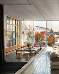 A modern living room, bathed in sunlight and featuring stylish décor, is showcased in "Ad At Home: Architectural Digest" by Random House. The space includes a beige sofa, cork stools, coffee table, art, and a hanging chair overlooking the river.