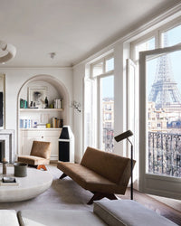 A modern living room, featured in Ad At Home: Architectural Digest by Random House, boasts brown furniture, large windows with Eiffel Tower views, built-in shelves filled with stylish décor, and sunlight highlighting thoughtful design.