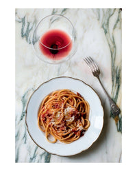 A plate of spaghetti inspired by Old World Italian from Random House, topped with tomato sauce and cheese, sits beside a glass of red wine and a fork on a marble surface.