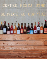 A row of colorful Salad Pizza Wine bottles by Random House stands on a wooden counter, set against a concrete wall with faded COFFEE PIZZA WINE SERVICE AU COMPTOIR text.