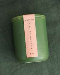 The LEIF Grapefruit & Pink Pepper Candle, a 4 oz green jar with a pink label, features a soy wax blend and wooden wick, and is shown resting on green fabric.
