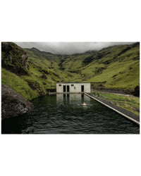 A person relaxes in the soothing waters of Hot Springs by Random House, swimming in a rectangular outdoor pool next to a small white building, surrounded by lush hills and mountains under a cloudy sky.