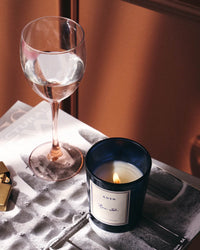 A ROEN Rue 52 Candle in a blue glass holder and a glass of clear liquid rest on a magazine, with a lighter nearby on a small table.