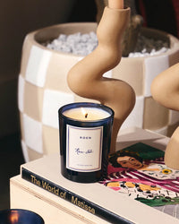 A lit ROEN Rue 52 Candle in Smoked Amber rests atop art books like The World of Matisse, beside a beige sculptural candleholder, with a checkered planter in the background.