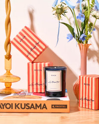 A black ROEN La Maison Rose Candle made from coconut soy wax sits on a Yayoi Kusama book, surrounded by red-striped boxes, a gold candle holder, and a vase with blue and white flowers.