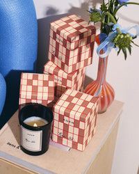 A ROEN Rue 52 Candle sits next to four stacked red and cream checkered boxes and a vase with blue flowers, all arranged on a book on a wooden surface.