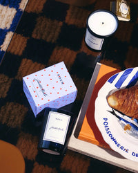 A ROEN Saint Malo Candle in glass with its box, a lit coconut soy wax candle, a croissant on a plate, and a closed book are arranged on a patterned table.