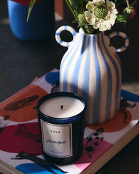 A blue and white striped vase with flowers, a ROEN Saint Malo Candle made from coconut soy wax with premium fragrance oils, and a colorful illustrated book are arranged on a table.