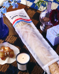 A ROEN Saint Malo Candle glows on the table alongside a baguette in a white cloth bag, pastries on a plate, fresh flowers in vases, and a decorative box.
