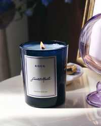 A lit ROEN Saint Malo Candle in blue glass, made with coconut soy wax and premium fragrance oils, rests on a white surface beside a purple glass piece and a small dish.