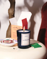 A lit ROEN La Maison Rose Candle, made from coconut soy wax with damask rose scent, rests in a dark glass jar on a side table near its open box, a matchbook, a matchstick, and a dish containing a tea bag packet.