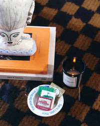 A stack of books with a ceramic head, a plate with matchboxes, and ROEN’s La Maison Rose Candle made from coconut soy wax rest on a checkered surface.