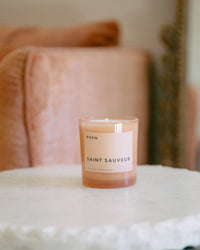 A peach-toned ROEN Saint Sauveur Candle sits on a round white marble table, with a plush chair and ornate gold decor softly blurred in the background.