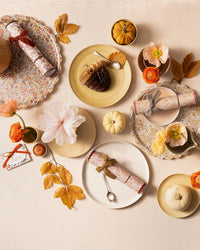 A festive autumn table features Liberty floral print plates, mini pumpkins, orange flowers, autumn leaves, spoons, a “happy” card, and Meri Meri x Liberty Fall Crackers with Velvet Bows—all atop a cream tablecloth.