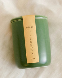 The LEIF Citrus & Oakmoss Candle, made from a soy wax blend and weighing 4 oz, comes in a green jar with a beige label and is shown resting on a light-colored textured surface.