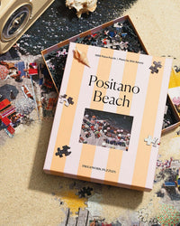 A Piecework Puzzles Positano Beach - Slim Aarons Collab 1000 Piece Puzzle in a pastel orange and cream striped box rests on sand, partially atop a brightly colored, half-assembled Amalfi Coast puzzle scene.