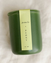 A LEIF Pomelo & Peony Candle, 4 oz, with a green jar, light green label, soy wax blend, and wooden wick is displayed on a light fabric surface.