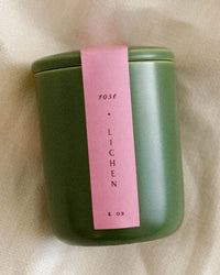 The LEIF Rose & Lichen Candle is a green ceramic jar with a pink label, 4 oz of soy wax. Stylishly displayed on light fabric, this mini candle adds elegant charm to any space.