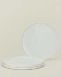Four Simple Porcelain Dinner Plates by Hawkins New York are displayed on a light beige background—three stacked horizontally and one propped vertically behind—highlighting their elegant fine porcelain style.