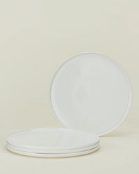 Four Simple Porcelain Dinnerware plates from Hawkins New York are displayed on a neutral background; three are stacked and one stands upright behind, highlighting this minimalist 16-piece set.