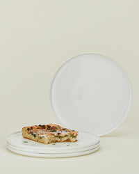 A stack of three Simple Porcelain Dinner Plates from Hawkins New York holds a rectangular slice of pizza, while a fourth plate from the set of 4 is propped upright behind them against a light beige background.