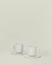 Two Simple Porcelain Espresso Cups & Saucers by Hawkins New York rest on a neutral background, their minimalist design and fine porcelain elegance enhanced by the surrounding empty space. Set includes 2 cups and saucers.