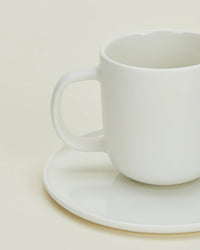 A Simple Porcelain Espresso Cup & Saucer from Hawkins New York sits empty and slightly off-center, its handle on the left, against a light cream background—showcasing minimalist elegance from this stylish set of 2.